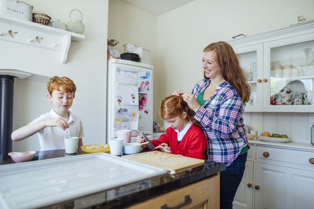 A medium shot of two redhead, Caucasian siblings having breakfast, the mother is helping plait her daughter's hair. They are both wearing school uniforms as it is their first day at school.